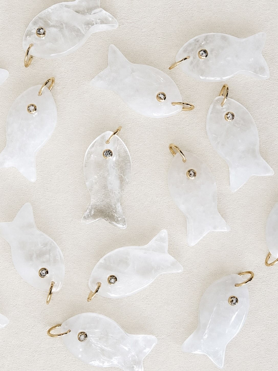 A flat-lay of multiple translucent white fish-shaped jewelry charms with small gold jump rings and tiny dark bead eyes, scattered on a light beige textured background.